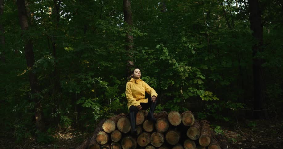 Woman wearing yellow raincoat sits on stack of logs among trees
