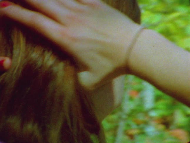 A girl is smiling and pulls back her hair as she holds on to her backpack and walks through the woods