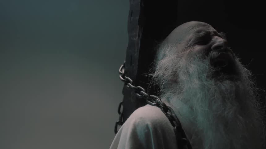 A bald man with a long white beard is chained to a wooden pole while screaming