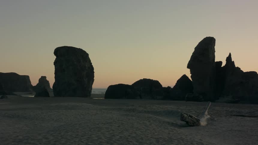Irregularly shaped driftwood sit on sandy beach a distance from rock outcroppings silhouetted against the red sky