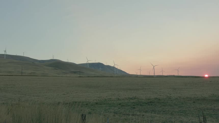 Wind turbines sit on hills near field with sun turning sky red from the horizon