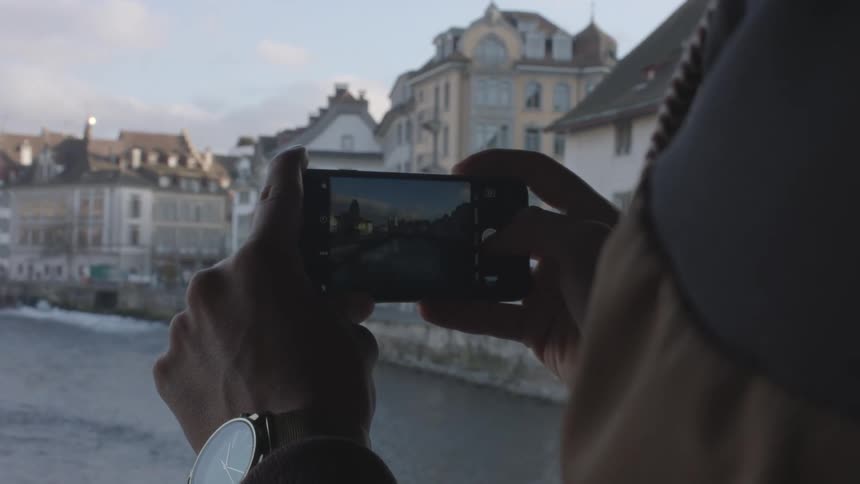 A person uses a smartphone to take photographs of a waterway in a city during the day