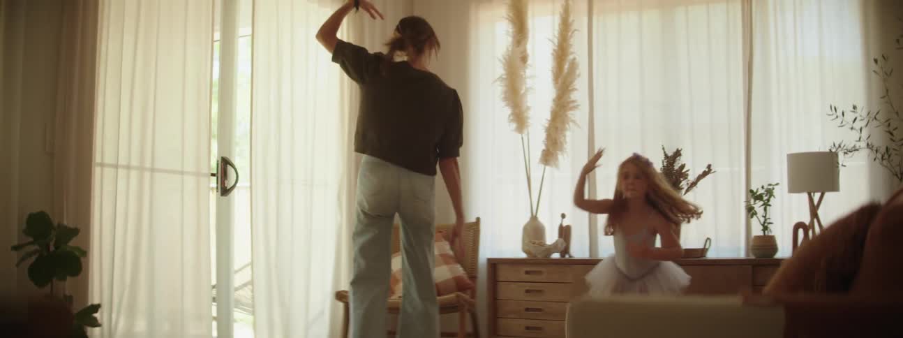 Mother with ponytail gray t shirt and ripped jeans dances with her daughter with red hair and tutu dances in the living room