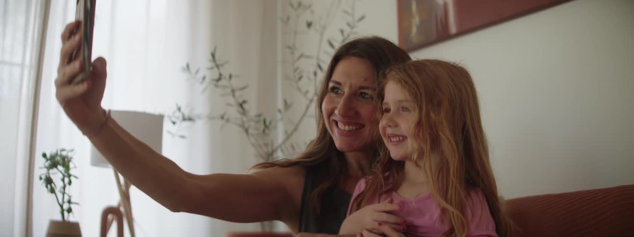 A little girl sits on her mothers lap at home while her mom hold a cell phone and takes a selfie of them