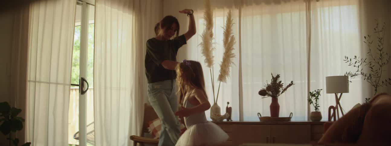 Mother with ponytail shirt and ripped jeans dances with her daughter with red hair and tutu in the living room