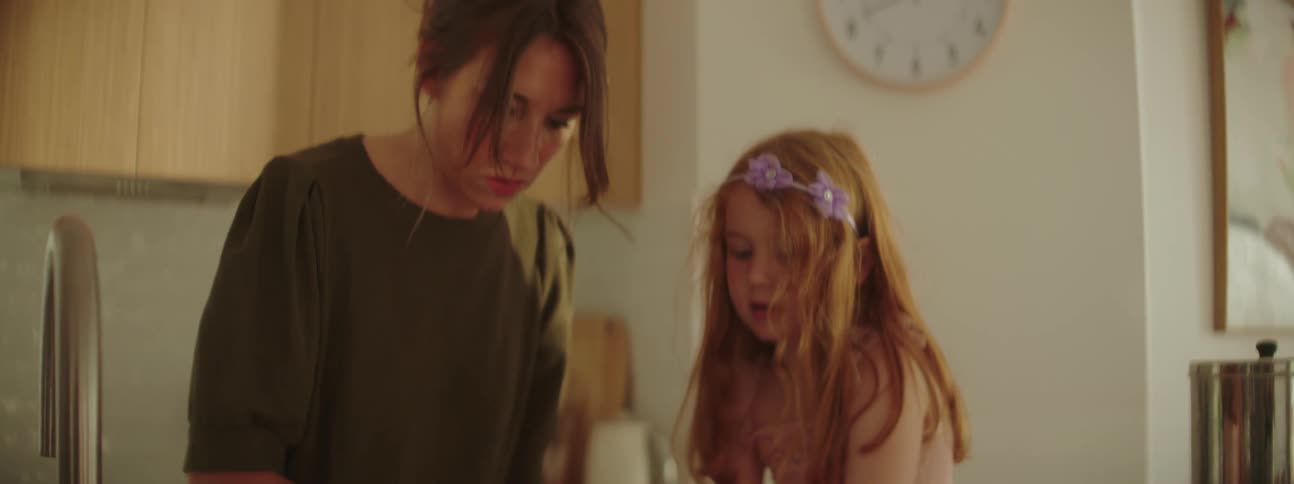 Mother with green blouse kneads dough on kitchen island next to daughter with red hair and pink dress in the kitchen