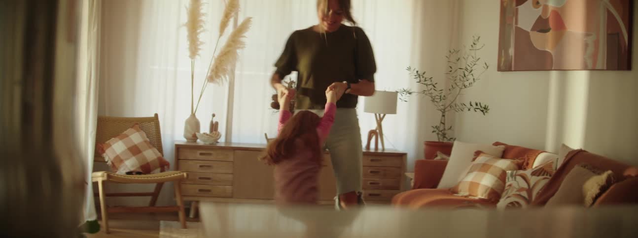 A woman is holding her young daughters hands and swinging her around in the living room