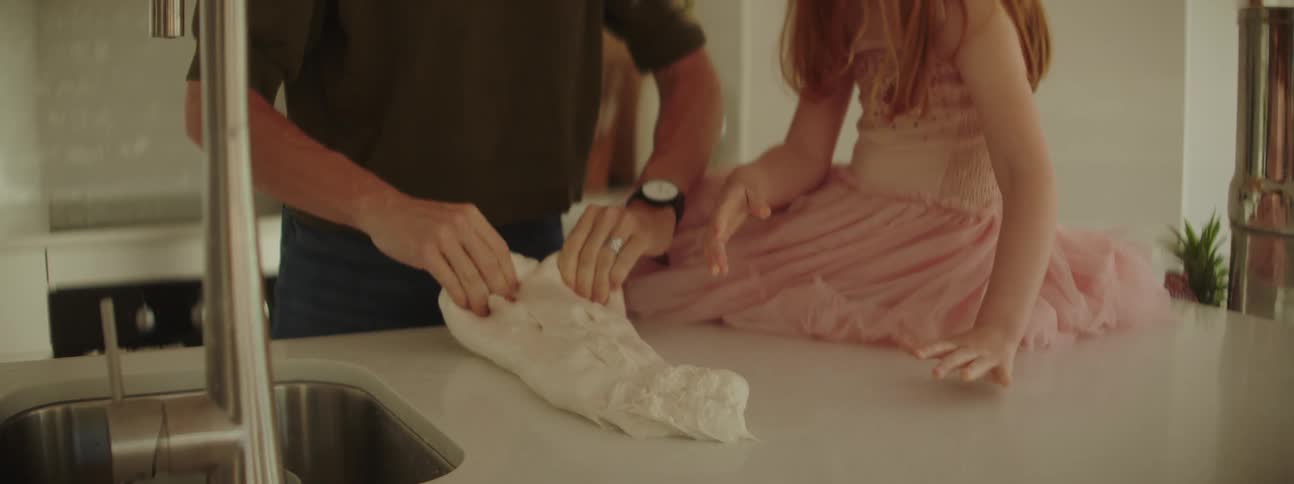 A woman is standing at the kitchen counter kneading dough and her little girl is sitting on the counter helping her