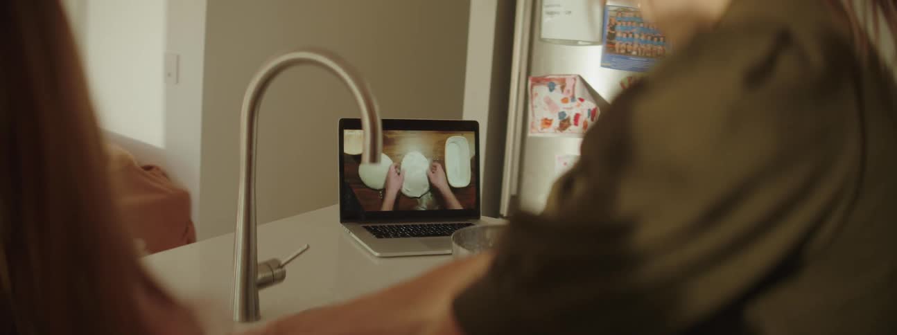 A little girl is in the kitchen with her mother with mixing bowls on the counter and a cooking video playing on the laptop computer