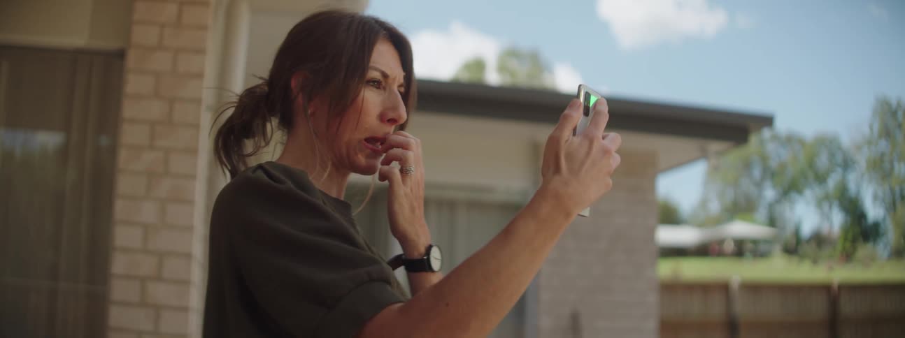 Woman with brown ponytail and dark green shirt makes video call on smartphone near the house in the backyard