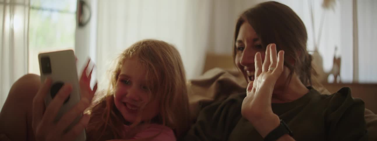 Women holding a phone waving and smiling with a little girl next to her smiling and waving