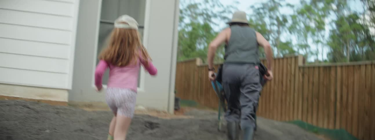 A woman is pushing a wheelbarrow to the back yard and her young daughter is running behind her