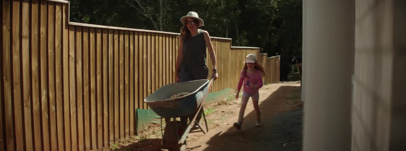 Woman with hat sunglasses gray top and pants pushes wheelbarrow next to daughter in the garden