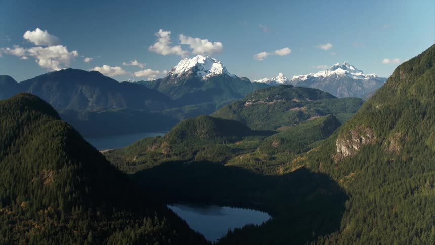 Aerial view of distant tall snowy mountains behind lakes and thick forested mountains