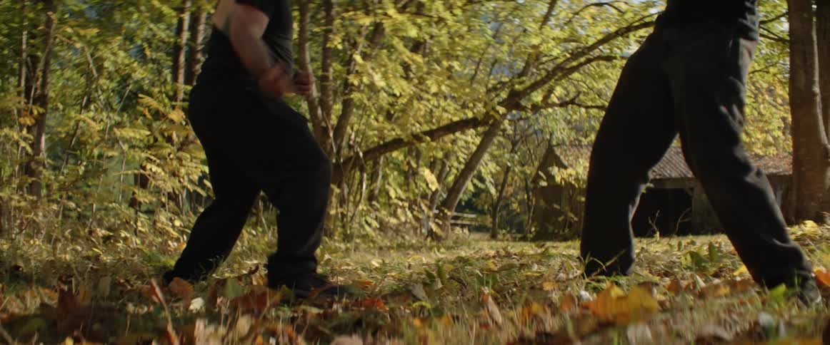 Two men practicing stick fighting on the grass near a cabin in the woods