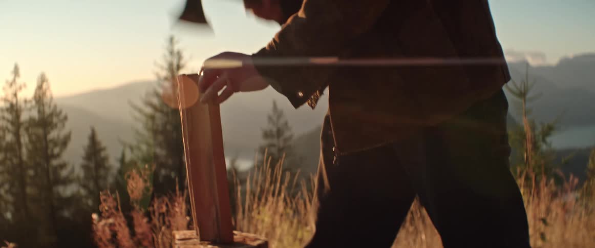 A man splits a piece of wood with a small axe in a mountain clearing at sunset
