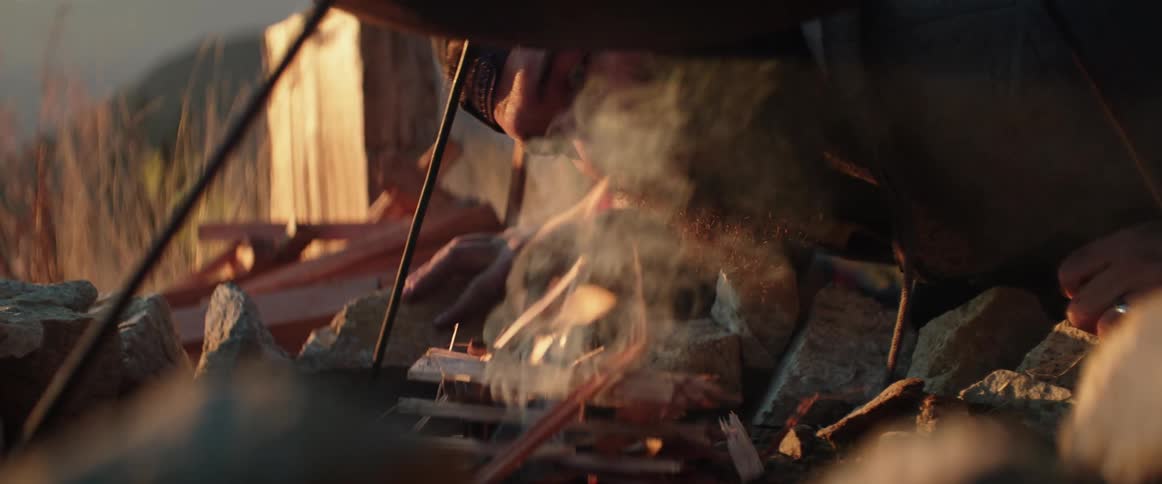 A man starts a fire underneath a cooking grill in a mountain clearing