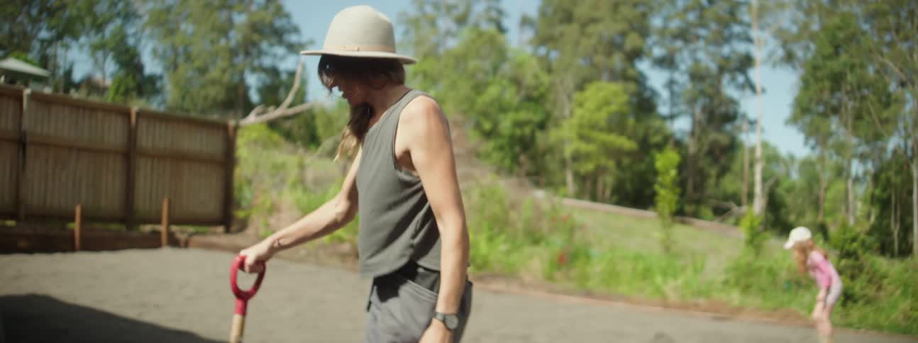 In backyard mother shovels dirt into wheelbarrow as daughter plays in the background