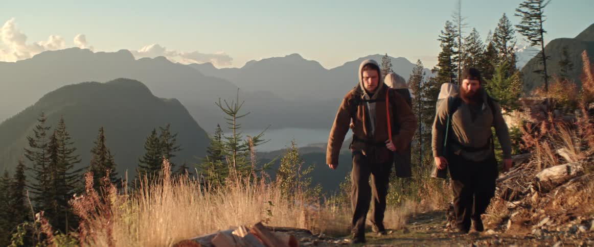 Two men wearing backpacks hike up a hill with distant mountains and they stop and take off their backpacks