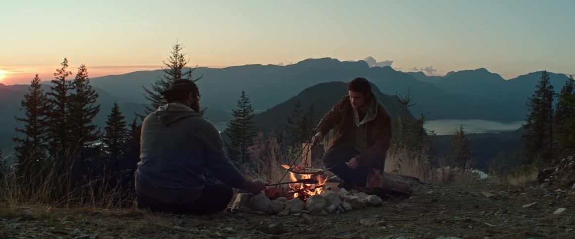 Two men are cooking by a campfire in the mountains at dusk