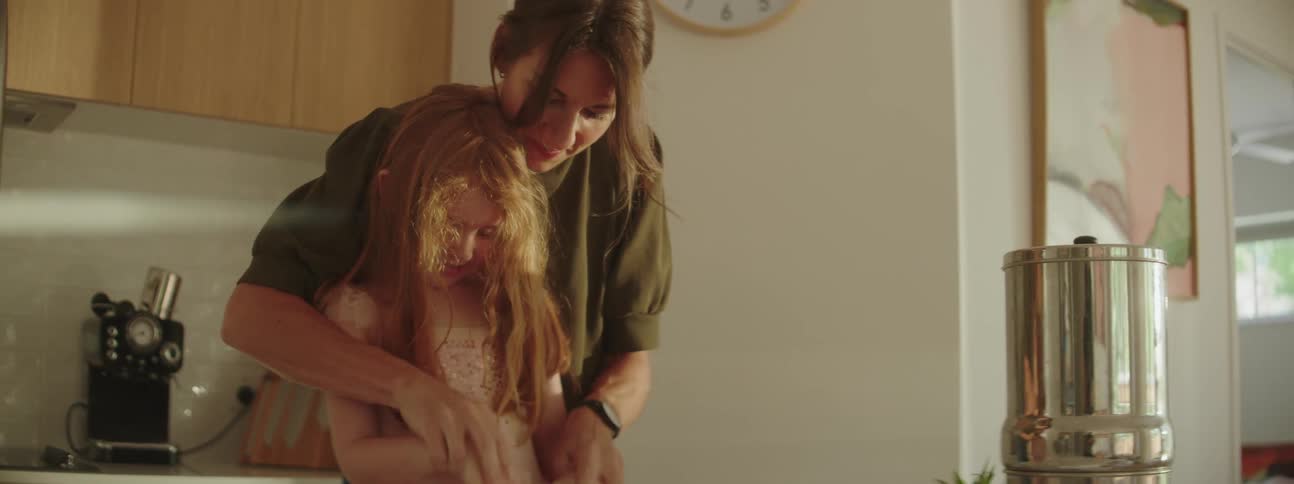 A mother playfully helps her daughter to knead dough in a home kitchen during the day