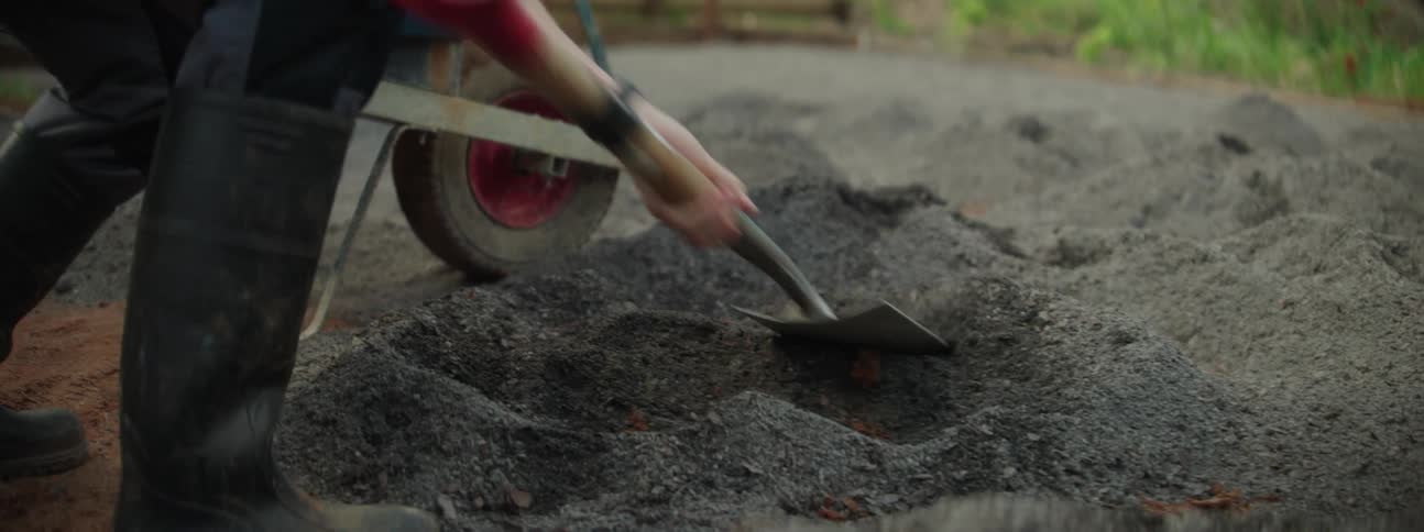 Person with black boot shovels soil in wheelbarrow