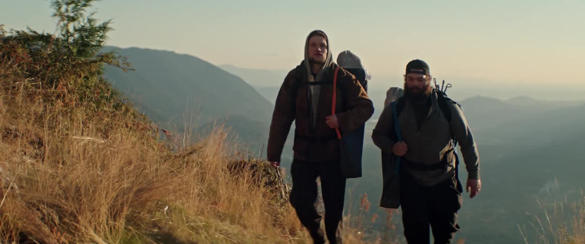 Two men wearing backpacks hike up a mountain trail from a valley