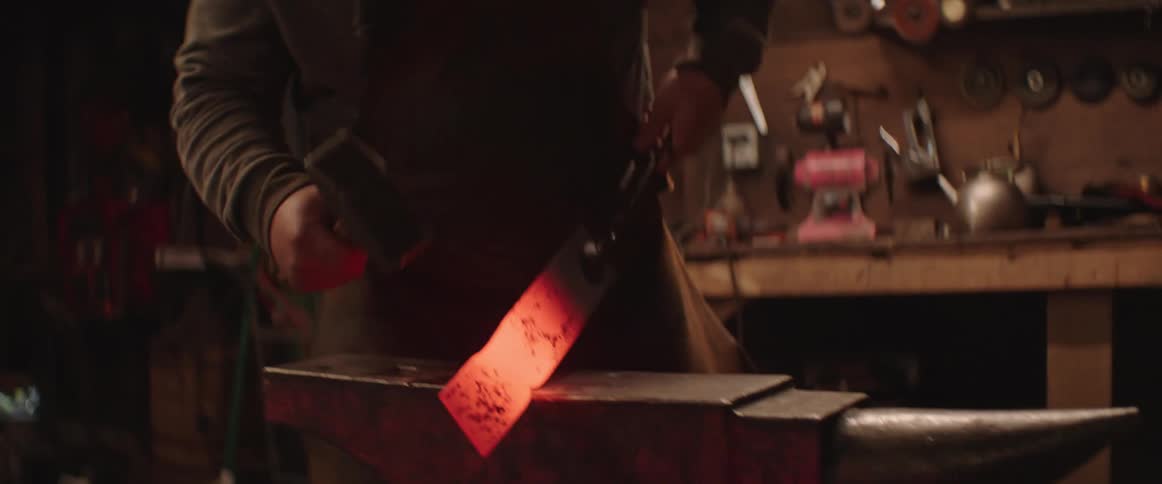 A blacksmith pulls a red hot piece of metal from a forge and begins to hammer it over an anvil