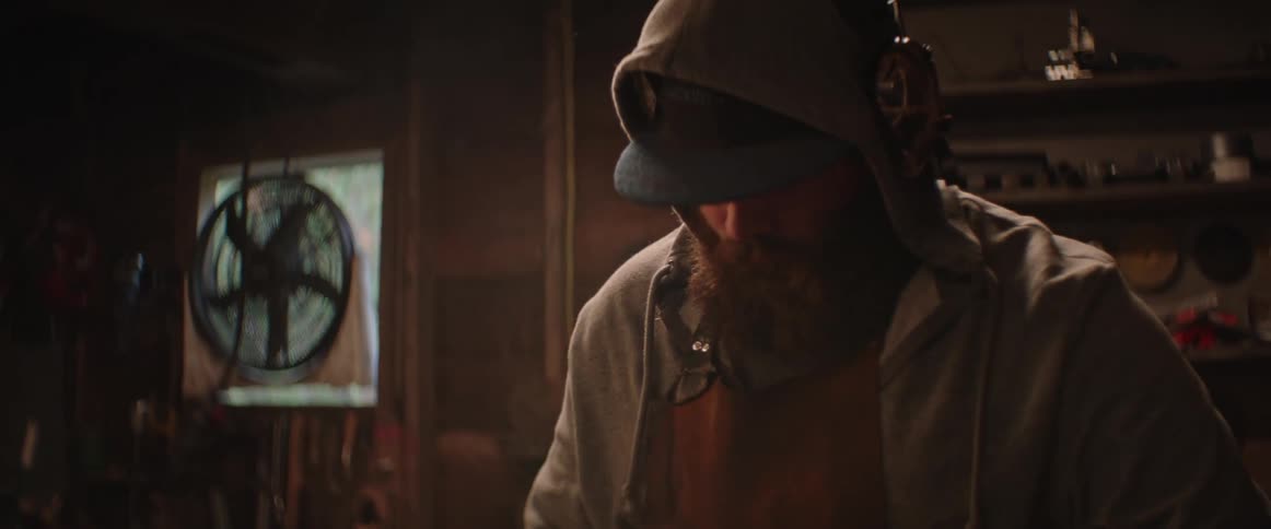 Bearded blacksmith wearing a hat hammering iron over an anvil