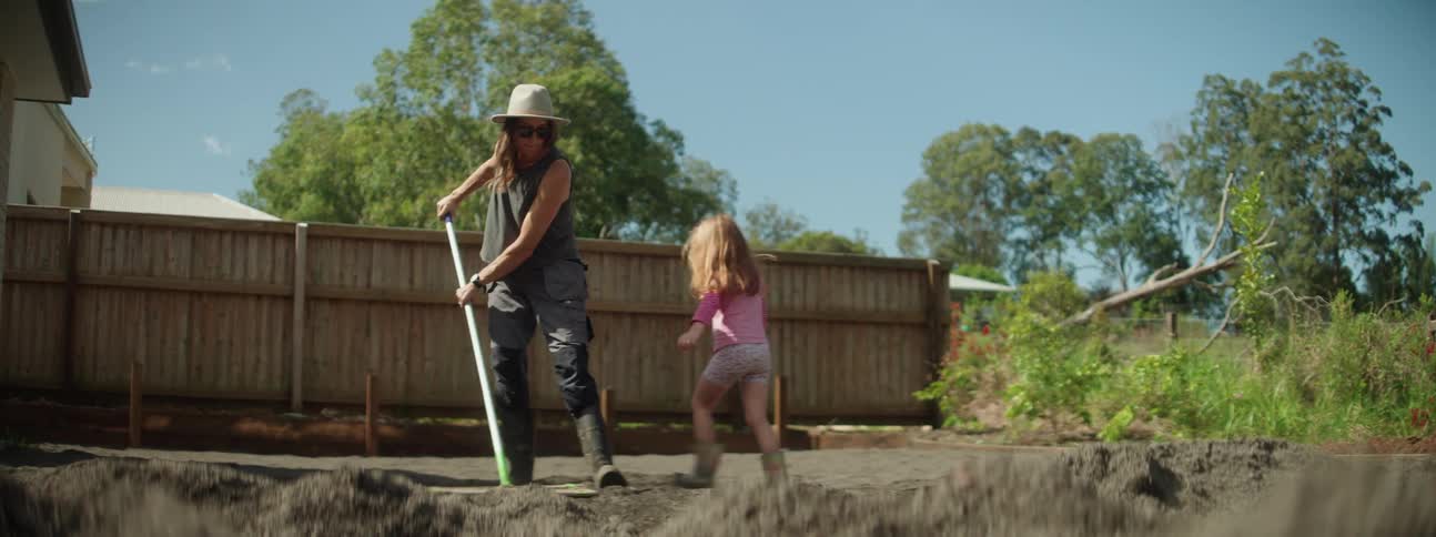 A mother rakes dirt while her daughter jumps and walks around her