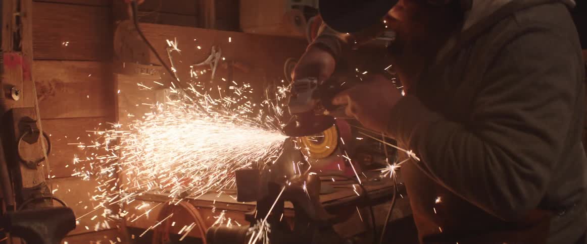 Worker with black hat and beard uses angle grinders to polish metal producing sparks