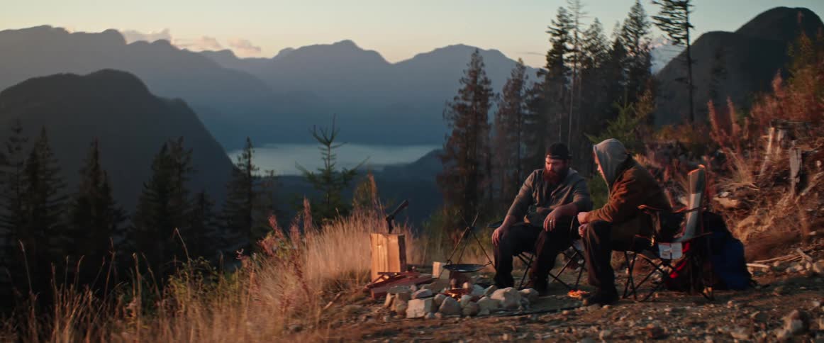 Two friends sitting in front of a campfire in a mountain clearing with a distant lake seen