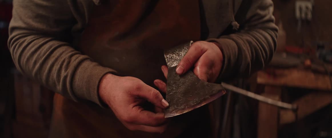 Man with brown leather apron and gray sweatshirt checks axe he made