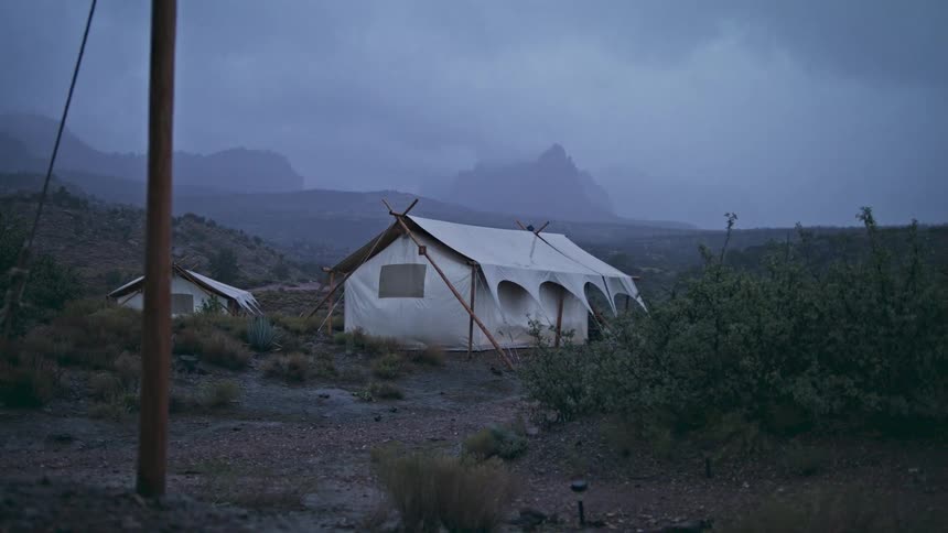 Two very large white tents are pitched in an area with just dirt grass and plants and the wind is blowing