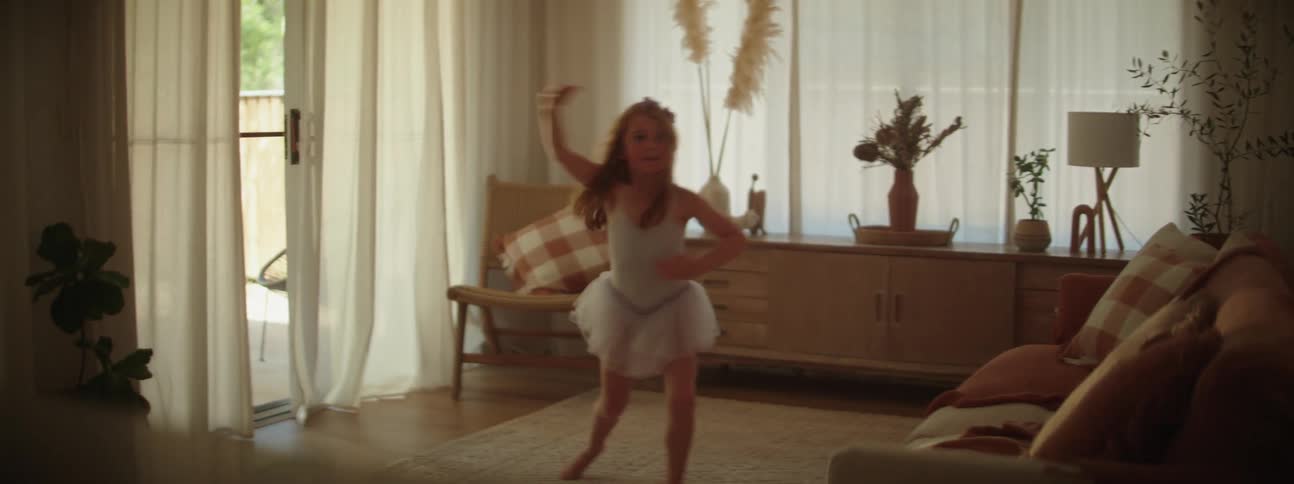 A girl dressed as a ballerina performs a clumsy ballet dance in the living room of a house during the day