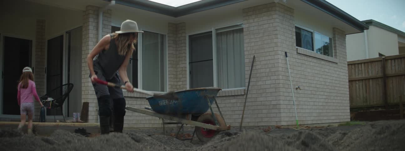Women shoveling dirt into a blue wheelbarrow and a little girl sitting on the step behind her