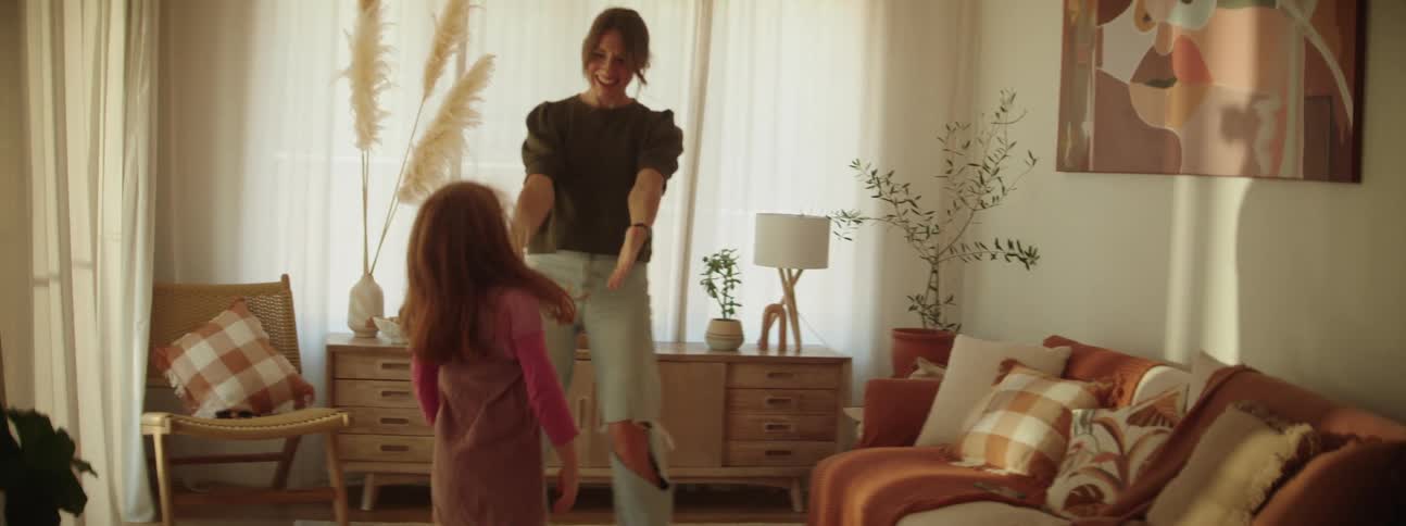 Woman with ponytail gray t shirt and ripped jeans dances with daughter with red hair in the living room