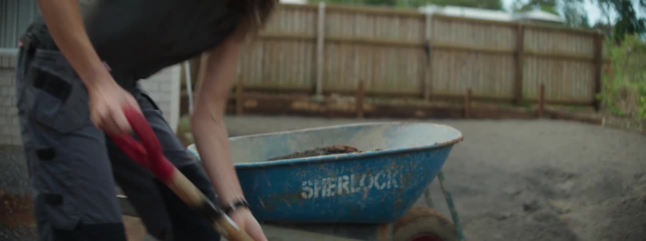 Woman wearing hat gray top and pants shovels sand in wheelbarrow in the garden