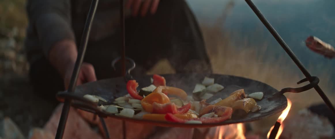 A person grills vegetables on a skillet over an open flame in the mountains