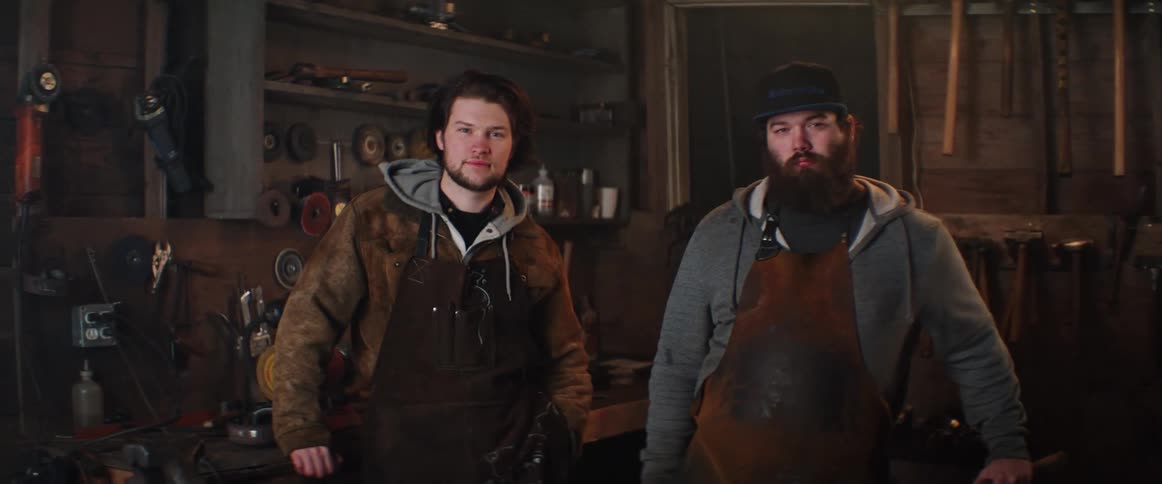 A pair of men wearing leather aprons look into the camera while standing together in a workshop