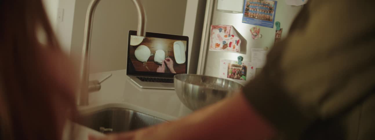 Two people watch cooking video on laptop while one follows recipe shown