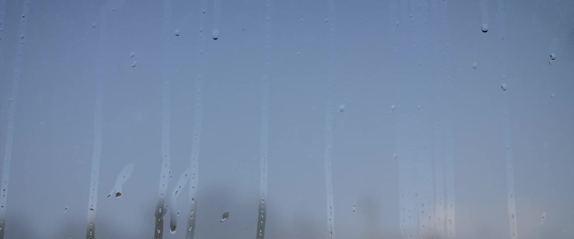 Water droplets are dripping down a steamed window and leave a trail as they move