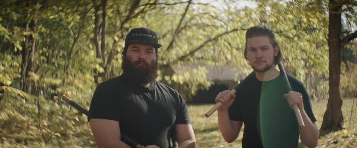 Two men stand in an autumn field holding tools with house in the background