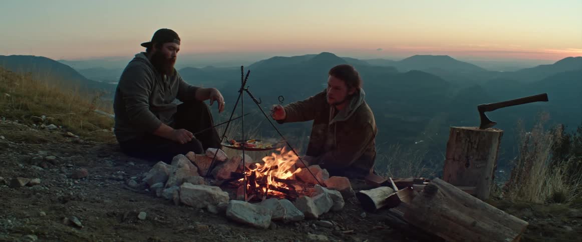 Two friends camping in the mountains cook food over a campfire at dusk