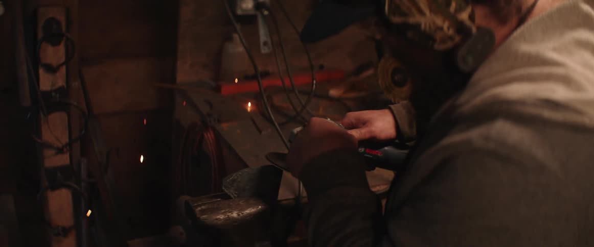 Worker with beard uses angle grinder to sharpen axe edge causing sparks