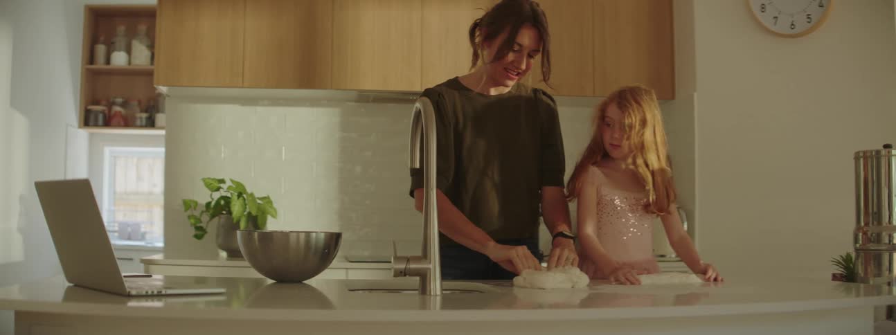 Mother with brown ponytail dark green blouse and daughter in pink tutu knead dough in the kitchen