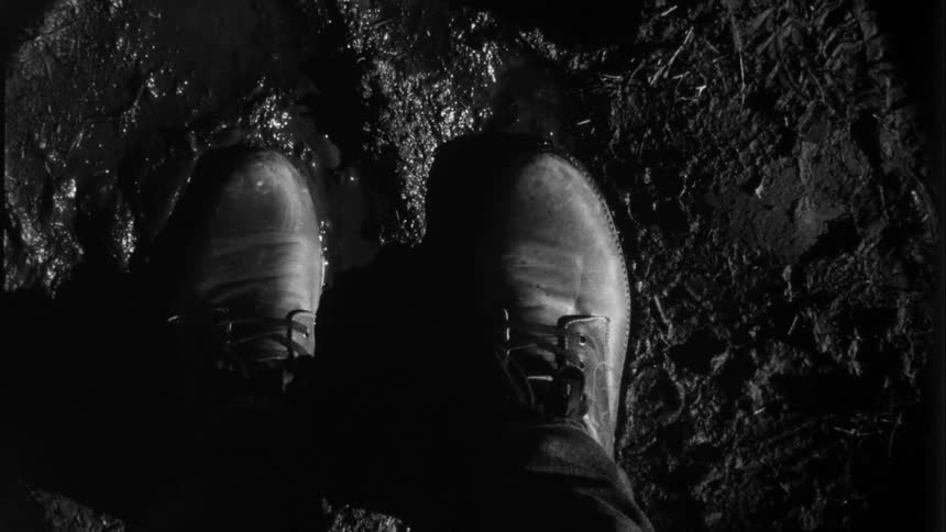 A person wearing boots are moving their feet in the mud as they stand in the rain at night and lightning flashes