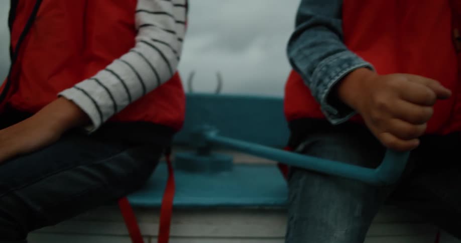 Two boys wearing red vests and jeans uses tiller to steer the boat