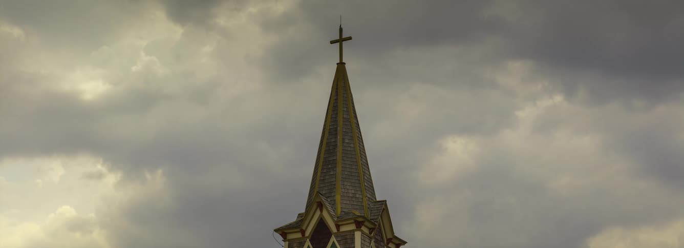 Gray clouds move over narrow tipped church roof topped with a cross