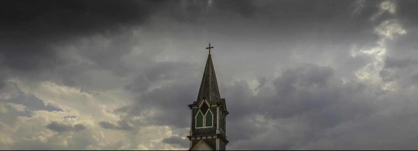 A cross stands tall on top of a church as dark clouds move across the sky